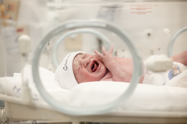 A newborn baby is crying inside an incubator wearing a white hat. The environment appears to be a hospital or neonatal unit, with medical equipment visible and a soft blanket on the incubator bed.