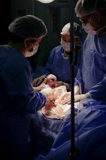 A newborn baby is being delivered in a hospital setting. Medical professionals wearing surgical gowns, masks, and hairnets assist in the delivery process. The infant is held gently, still covered in bodily fluids, against a backdrop of surgical drapes and equipment.
