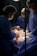 A newborn baby is being delivered in a hospital setting. Medical professionals wearing surgical gowns, masks, and hairnets assist in the delivery process. The infant is held gently, still covered in bodily fluids, against a backdrop of surgical drapes and equipment.