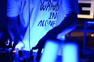 A person wearing a white T-shirt with text, standing in a dimly lit environment and engaging with a DJ mixer. The atmosphere is illuminated by blue light, creating a vibrant and energetic ambiance.