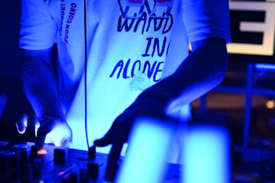 A person wearing a white T-shirt with text, standing in a dimly lit environment and engaging with a DJ mixer. The atmosphere is illuminated by blue light, creating a vibrant and energetic ambiance.