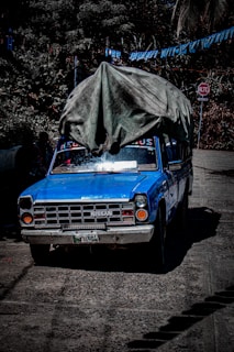 A blue Nissan pickup truck covered with a large, worn-out tarp is parked on a textured road. Surrounding the vehicle is lush greenery with dense foliage, and above it, blue and white flags are strung across. A stop sign with the word 'Alto' is visible in the background.