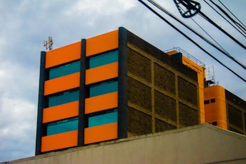 A multi-story building with a modern design, featuring bright orange rectangular panels and dark-tinted windows. The structure has industrial elements, including visible wiring and antennas on the rooftop. The facade is made of a mix of orange and brown brick materials, conveying an urban aesthetic against a cloudy sky backdrop.