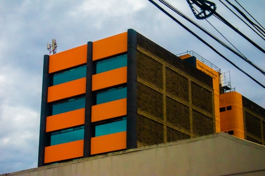 A multi-story building with a modern design, featuring bright orange rectangular panels and dark-tinted windows. The structure has industrial elements, including visible wiring and antennas on the rooftop. The facade is made of a mix of orange and brown brick materials, conveying an urban aesthetic against a cloudy sky backdrop.