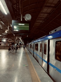 A train station platform featuring a clock showing the time as 22:06. The platform is well-lit, with a train on the right side and several people walking or standing near the tracks. An electronic sign displays the destination as 'Airport'. The interior architecture is marked by a high, arched metallic ceiling.