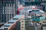 An urban landscape featuring a mix of modern and traditional architecture. Several buildings showcase a combination of brickwork and glass. A red building stands out amidst the predominantly muted tones. Rooftops exhibit various geometric designs, and there is visible greenery in the background.