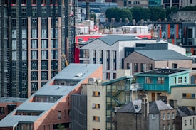 An urban landscape featuring a mix of modern and traditional architecture. Several buildings showcase a combination of brickwork and glass. A red building stands out amidst the predominantly muted tones. Rooftops exhibit various geometric designs, and there is visible greenery in the background.