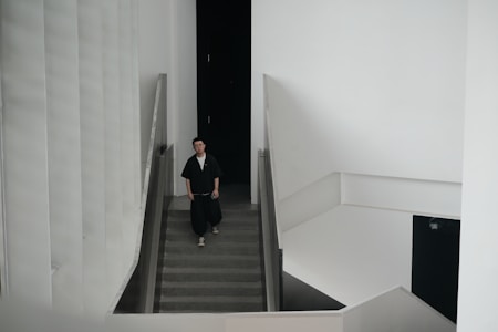 A person dressed in dark clothing is walking down a staircase in a minimalistic, modern building with white walls and clean architectural lines. The building has a high ceiling and features a large, black door at the top of the stairs.