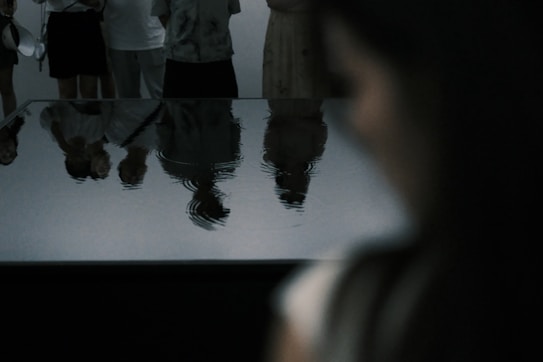 A group of people is reflected in a water surface, creating distorted, ripple-like textures. The scene is dimly lit, suggesting an indoor setting with an artistic or contemplative atmosphere. The focus is on the patterns created by the reflections.