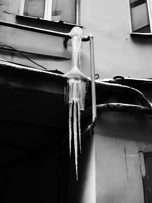 A large icicle extends down from a pipeline on the side of a building. The icicle is formed from frozen water dripping from the pipes above. Part of the building's facade, including windows and the pipe structure, is visible in the background.