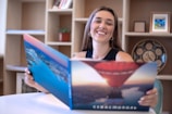 A woman with long hair is smiling while holding a large open book or brochure with images of a hot air balloon and a river. She is seated at a white table against a background of shelves containing books, a decorative plate, and framed pictures.