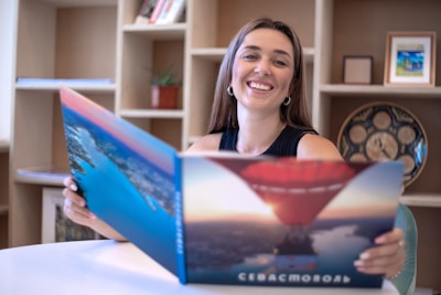 A woman with long hair is smiling while holding a large open book or brochure with images of a hot air balloon and a river. She is seated at a white table against a background of shelves containing books, a decorative plate, and framed pictures.