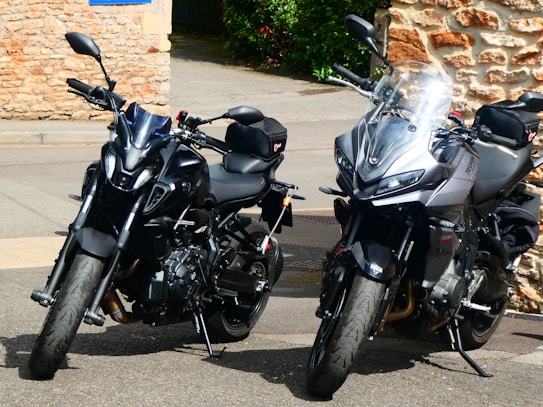 Two sleek, modern motorcycles are parked on a street lined with light-colored bricks and a green bush in the background. The motorcycles are well-maintained with shiny black and grey metallic finishes, featuring deep-tread tires and multiple storage compartments.