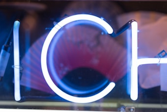 A close-up of illuminated neon signs featuring the letters I, C, and H. The neon is bright blue against a darker background with some red and orange hues faintly visible.
