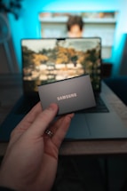 A hand holds a portable Samsung solid-state drive in front of an open laptop. In the background, a blurred image of a person using the laptop is visible, with a brightly lit display showing a scenic wallpaper.