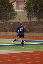 A soccer player wearing a dark blue uniform is running on a field with artificial turf while light snow is falling. Their back is facing the camera, showcasing the number 17 on the jersey. The field has red and green sections, typical of a sports complex, and there are trees and a fence in the background.