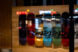 Multiple skateboard decks displayed vertically, each printed with the same image of an electric chair in varying color schemes. The image is reflected against a glass surface, creating a layered visual effect. The artwork is attributed to Andy Warhol, blending pop art themes with skate culture.