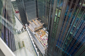 A construction site located between tall modern skyscrapers is visible from an elevated perspective. The buildings are primarily made of glass and steel, and the site includes scaffolding and construction materials. A red double-decker bus can be seen in the background on the street.