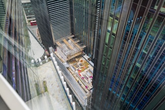 A construction site located between tall modern skyscrapers is visible from an elevated perspective. The buildings are primarily made of glass and steel, and the site includes scaffolding and construction materials. A red double-decker bus can be seen in the background on the street.