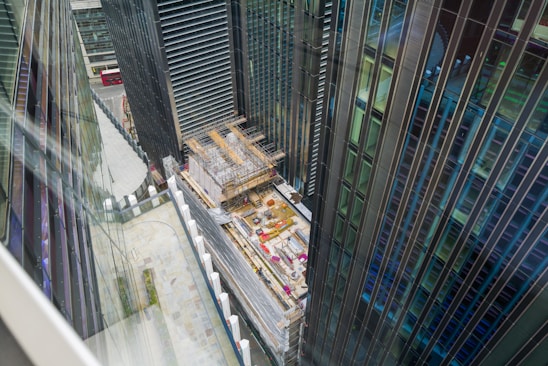 A construction site located between tall modern skyscrapers is visible from an elevated perspective. The buildings are primarily made of glass and steel, and the site includes scaffolding and construction materials. A red double-decker bus can be seen in the background on the street.