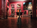 An art gallery interior with several large, classical paintings displayed on deep red walls. The floor is made of polished wood with decorative patterns. Ornate, classical-style furniture and vases are placed around the room. The lighting is soft, highlighting the artwork.