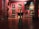 An art gallery interior with several large, classical paintings displayed on deep red walls. The floor is made of polished wood with decorative patterns. Ornate, classical-style furniture and vases are placed around the room. The lighting is soft, highlighting the artwork.