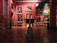 An art gallery interior with several large, classical paintings displayed on deep red walls. The floor is made of polished wood with decorative patterns. Ornate, classical-style furniture and vases are placed around the room. The lighting is soft, highlighting the artwork.