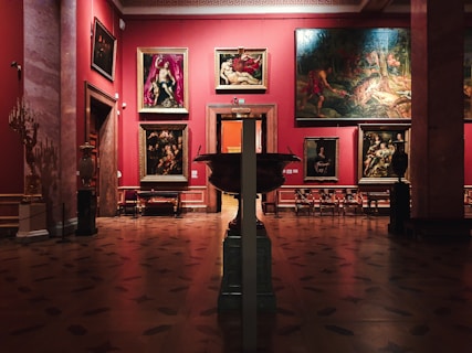 An art gallery interior with several large, classical paintings displayed on deep red walls. The floor is made of polished wood with decorative patterns. Ornate, classical-style furniture and vases are placed around the room. The lighting is soft, highlighting the artwork.