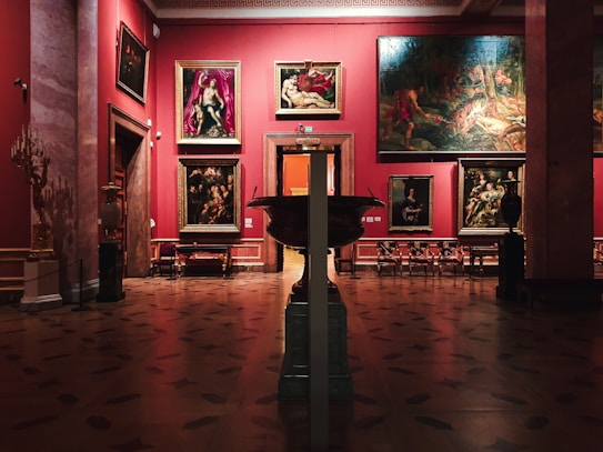 An art gallery interior with several large, classical paintings displayed on deep red walls. The floor is made of polished wood with decorative patterns. Ornate, classical-style furniture and vases are placed around the room. The lighting is soft, highlighting the artwork.