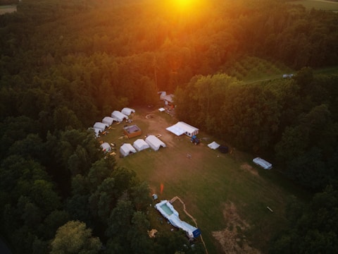 An aerial view of a campsite surrounded by dense forest, featuring several white tents arranged in an open clearing. The sun is setting in the background, casting a warm golden light over the scene. There are various structures and pathways visible among the tents.