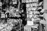 A small shop is filled with a variety of household items and daily necessities. Products are densely packed on shelves and hangers, including bags, kitchen utensils, electronic accessories, toys, and cleaning supplies. A person wearing a hat is sitting inside the shop, partially hidden by the merchandise. The scene is captured in black and white, adding a nostalgic feel to the cluttered but organized store setting.