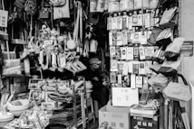 A small shop is filled with a variety of household items and daily necessities. Products are densely packed on shelves and hangers, including bags, kitchen utensils, electronic accessories, toys, and cleaning supplies. A person wearing a hat is sitting inside the shop, partially hidden by the merchandise. The scene is captured in black and white, adding a nostalgic feel to the cluttered but organized store setting.