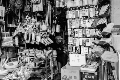 A small shop is filled with a variety of household items and daily necessities. Products are densely packed on shelves and hangers, including bags, kitchen utensils, electronic accessories, toys, and cleaning supplies. A person wearing a hat is sitting inside the shop, partially hidden by the merchandise. The scene is captured in black and white, adding a nostalgic feel to the cluttered but organized store setting.