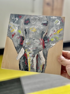 A hand holds a piece of artwork depicting an abstract elephant. The elephant's face is painted with bold brushstrokes in shades of gray, yellow, red, and white. The painting is on a rectangular wooden board, and the background appears to be an indoor setting with blurred elements.
