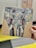 A hand holds a piece of artwork depicting an abstract elephant. The elephant's face is painted with bold brushstrokes in shades of gray, yellow, red, and white. The painting is on a rectangular wooden board, and the background appears to be an indoor setting with blurred elements.