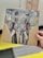 A hand holds a piece of artwork depicting an abstract elephant. The elephant's face is painted with bold brushstrokes in shades of gray, yellow, red, and white. The painting is on a rectangular wooden board, and the background appears to be an indoor setting with blurred elements.
