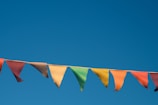 A string of colorful pennant flags is suspended against a clear blue sky. The flags are triangular and vary in colors including red, orange, yellow, green, and blue. There are small frays visible at the edges of some of the flags, indicating weathering or wear.