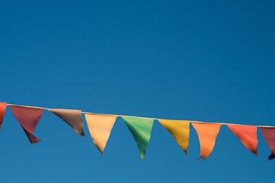 A string of colorful pennant flags is suspended against a clear blue sky. The flags are triangular and vary in colors including red, orange, yellow, green, and blue. There are small frays visible at the edges of some of the flags, indicating weathering or wear.