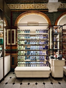 A display of neatly arranged colorful tins on shelving units within a store. The shelves hold tins in shades of blue, purple, and green, with a decorative sign above reading 'CHOCOLATE COATED'. The background features ornate architectural details with tiles and arches.