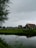 A picturesque rural landscape with traditional Dutch houses, characterized by their dark green and black exteriors with white trims. The scene includes a calm body of water in the foreground, reflecting the houses and the overcast sky above. Lush green grass surrounds the area, and a few people are seen walking near the houses, adding life to the tranquil setting. A wooden bridge crosses a small stream, enhancing the pastoral atmosphere.