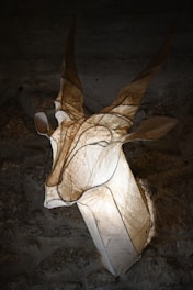 An artistic sculpture resembling a gazelle or antelope head is mounted on a textured stone wall. The sculpture appears to be made of a translucent material, allowing light to shine through and create a glowing effect. The details of the horns and ears are highlighted by the lighting, creating a dramatic and ethereal presence.