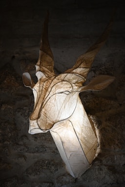 An artistic sculpture resembling a gazelle or antelope head is mounted on a textured stone wall. The sculpture appears to be made of a translucent material, allowing light to shine through and create a glowing effect. The details of the horns and ears are highlighted by the lighting, creating a dramatic and ethereal presence.