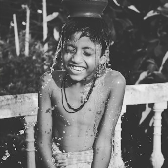 A child is joyfully playing with water pouring from a pot placed on their head. The expression is one of delight and laughter, with water droplets splashing around. The scene captures a moment of pure happiness.