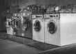 A row of industrial washing machines is lined up, each with a porthole door. Above the machines are displayed various laundry-related products, possibly for purchase. The machines have visible dials and instructions, suggesting they are coin-operated or commercial laundromat machines. The setting appears to be in a functional, practical environment with a tiled floor.