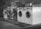A row of industrial washing machines is lined up, each with a porthole door. Above the machines are displayed various laundry-related products, possibly for purchase. The machines have visible dials and instructions, suggesting they are coin-operated or commercial laundromat machines. The setting appears to be in a functional, practical environment with a tiled floor.