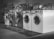 A row of industrial washing machines is lined up, each with a porthole door. Above the machines are displayed various laundry-related products, possibly for purchase. The machines have visible dials and instructions, suggesting they are coin-operated or commercial laundromat machines. The setting appears to be in a functional, practical environment with a tiled floor.