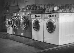A row of industrial washing machines is lined up, each with a porthole door. Above the machines are displayed various laundry-related products, possibly for purchase. The machines have visible dials and instructions, suggesting they are coin-operated or commercial laundromat machines. The setting appears to be in a functional, practical environment with a tiled floor.