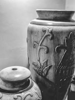 Two intricately designed ceramic vases are placed close together. The larger vase has floral carvings, while the smaller one has a lid and additional ornamental designs. The vases exhibit a rustic, handmade quality.