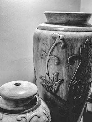 Two intricately designed ceramic vases are placed close together. The larger vase has floral carvings, while the smaller one has a lid and additional ornamental designs. The vases exhibit a rustic, handmade quality.