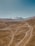 A vast and arid landscape extends towards rugged mountains capped with snow under a clear blue sky. The terrain is predominantly brown and barren, with winding dirt roads traversing through it. A small settlement can be seen in the middle of the plain, adding a hint of human presence in this natural setting.